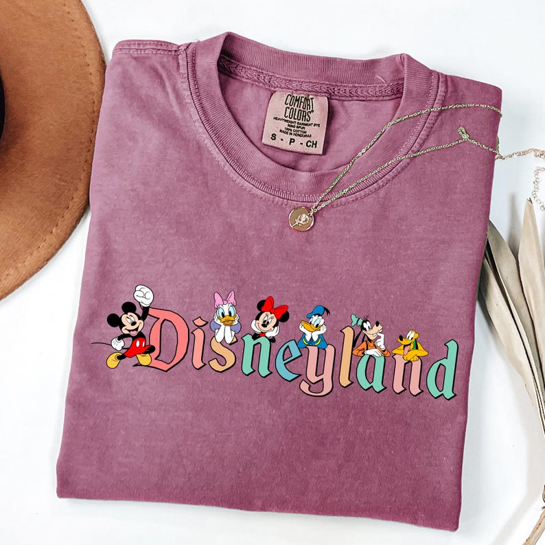 May include: A mauve-colored t-shirt with the word "Disneyland" in a colorful, stylized font. The shirt features cartoon characters Mickey Mouse, Daisy Duck, Minnie Mouse, Donald Duck, Goofy, and Pluto. A gold necklace is draped across the shirt.