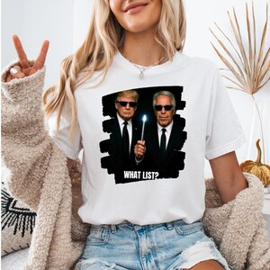 K&ouml;nnte beinhalten: Wei&szlig;es T-Shirt mit einer Grafik von zwei M&auml;nnern in schwarzen Anz&uuml;gen und Sonnenbrillen, einer h&auml;lt einen leuchtenden Zauberstab. Der Text "WHAT LIST?" befindet sich unter dem Bild. Das Shirt ist ein l&auml;ssiger Stil.