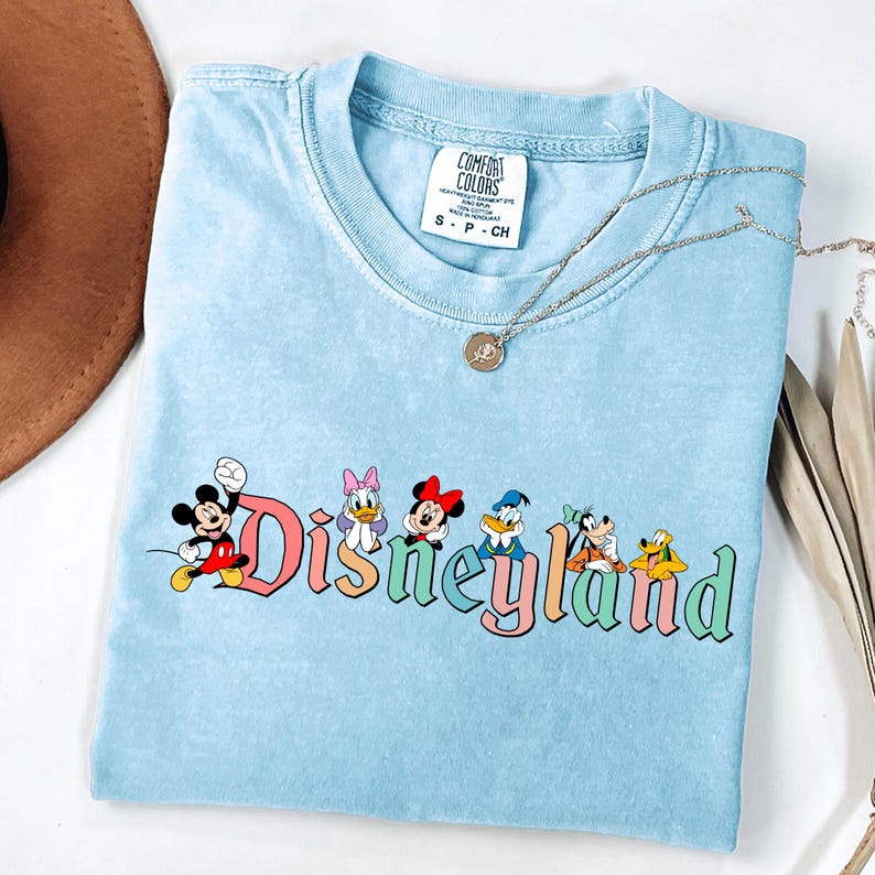 May include: Light blue t-shirt with the word "Disneyland" in colorful letters and cartoon characters. The shirt features Mickey Mouse, Daisy Duck, Minnie Mouse, Donald Duck, Goofy, and Pluto. A silver necklace with a pendant is on top of the shirt.