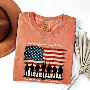 May include: A peach-colored t-shirt with a distressed American flag graphic and the text "land of the free, because of the brave".  Below the flag are seven silhouettes of soldiers with the text "honor and remember" on either side.