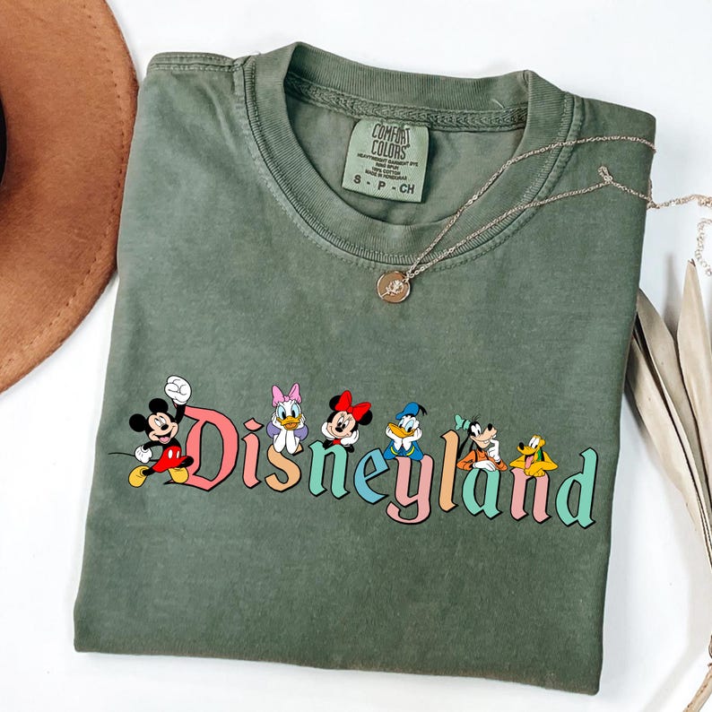 May include: Olive green t-shirt featuring the word "Disneyland" in colorful letters with cartoon characters. The shirt has a crew neck and short sleeves. A gold necklace with a pendant is draped across the shirt.