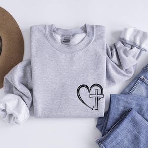 May include: A gray sweatshirt with a black graphic of a heart with a cross inside. The sweatshirt is a crew neck style.