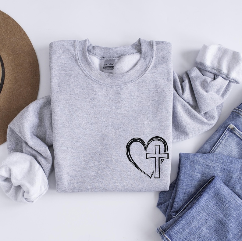 May include: A gray sweatshirt with a black graphic of a heart with a cross inside. The sweatshirt is a crew neck style.