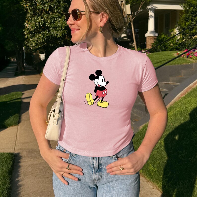 May include: A light pink t-shirt featuring a graphic of Mickey Mouse. The cartoon character is depicted in his classic outfit: black shorts, yellow shoes, and red shorts. The shirt is paired with blue jeans.