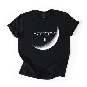 May include: Black t-shirt featuring the text "ARTEMIS II" in white, above a detailed crescent moon graphic. The shirt is made of a soft material and has rolled-up sleeves.