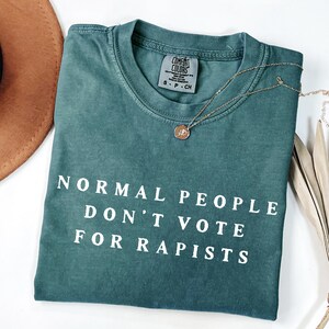 以下が含まれることがあります： 「NORMAL PEOPLE DON'T VOTE FOR RAPISTS」の白い文字が入った、ティール色のComfort Colors Tシャツ。ペンダント付きのゴールドネックレスがシャツの上にかけられています。茶色の帽子が左上に写っています。