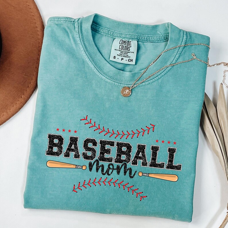 May include: A teal blue t-shirt with a distressed look featuring a baseball graphic with the text "Baseball mom" and two crossed baseball bats.