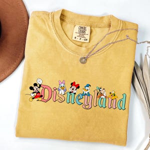 May include: Mustard yellow t-shirt with the word "Disneyland" in colorful letters and cartoon characters. The shirt has a crew neck and short sleeves. A silver necklace with a pendant is on top of the shirt.