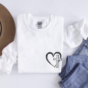 May include: White sweatshirt with a black and white graphic of a heart with a cross inside. The sweatshirt is laying on a white surface with a pair of blue jeans.