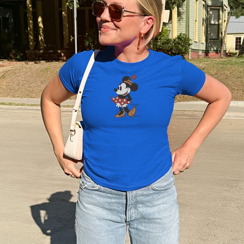 May include: A royal blue t-shirt featuring a graphic of Minnie Mouse in a polka dot dress and a festive hat. The cartoon character is depicted in a walking pose. The shirt is paired with light wash denim jeans.