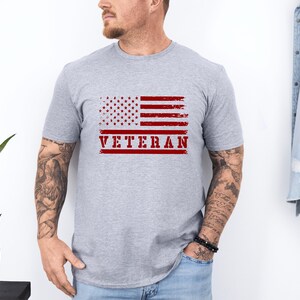 以下が含まれることがあります： 赤い文字で「VETERAN」と印刷された、くたびれたアメリカ国旗が描かれたグレーのTシャツです。