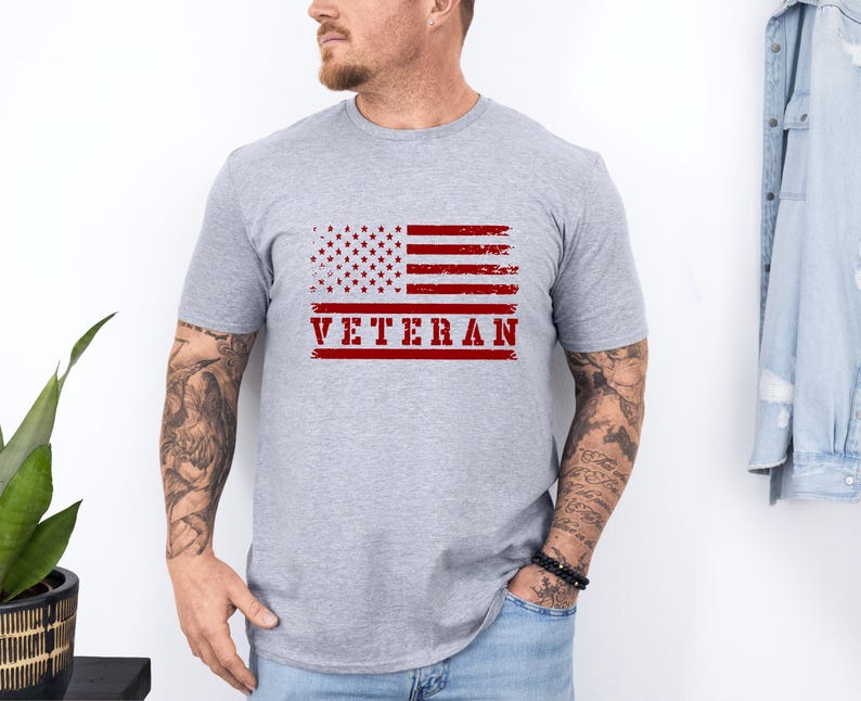 以下が含まれることがあります： 赤い文字で「VETERAN」と印刷された、くたびれたアメリカ国旗が描かれたグレーのTシャツです。