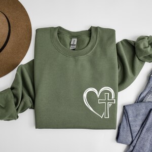 May include: A green sweatshirt with a white graphic of a heart with a cross inside. The sweatshirt is a crew neck style.