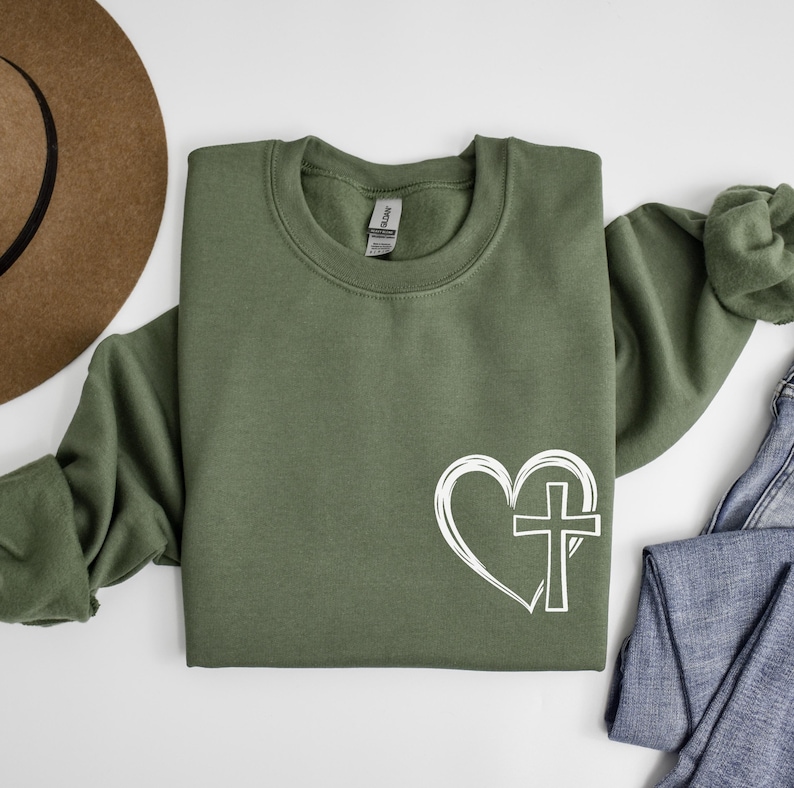 May include: A green sweatshirt with a white graphic of a heart with a cross inside. The sweatshirt is a crew neck style.
