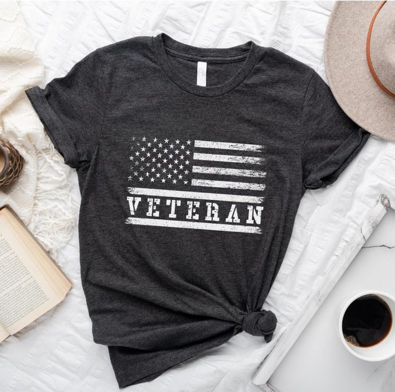 以下が含まれることがあります： くすんだグレーのTシャツに、白くぼやけたアメリカの旗と「VETERAN」の文字がプリントされています。