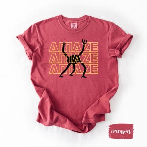 May include: Crimson-colored t-shirt with rolled-up sleeves. The shirt features the word "AMAZE" stacked three times in a yellow font, with a black stick figure design incorporated into the letters. The shirt has a crew neck.