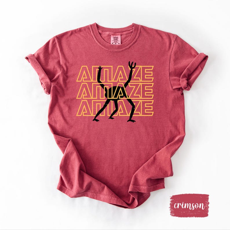 May include: Crimson-colored t-shirt with rolled-up sleeves. The shirt features the word "AMAZE" stacked three times in a yellow font, with a black stick figure design incorporated into the letters. The shirt has a crew neck.