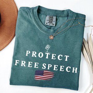 May include: A teal Comfort Colors t-shirt with the words "PROTECT FREE SPEECH" in white, along with a graphic of a microphone and a distressed American flag. A gold necklace with a pendant is draped on the shirt.