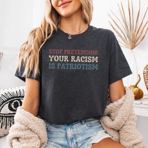 Pode incluir: Uma camiseta cinza escura mesclada com o texto "STOP PRETENDING YOUR RACISM IS PATRIOTISM" impresso em uma fonte de estilo retr&ocirc; com acentos vermelhos, azuis e amarelos.