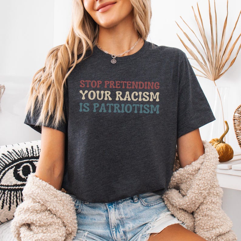Pode incluir: Uma camiseta cinza escura mesclada com o texto "STOP PRETENDING YOUR RACISM IS PATRIOTISM" impresso em uma fonte de estilo retr&ocirc; com acentos vermelhos, azuis e amarelos.