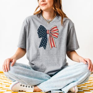 Peut inclure: Un t-shirt gris avec un n&oelig;ud rouge, blanc et bleu. Le n&oelig;ud est con&ccedil;u pour ressembler au drapeau am&eacute;ricain.
