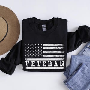 May include: Black sweatshirt with a white distressed American flag and the word "Veteran" printed below it.