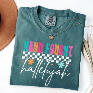 May include: A teal Comfort Colors t-shirt with the words "HARD FOUGHT hallelujah" in colorful text. The design includes a checkered pattern and floral accents. A gold necklace with a pendant is draped on the shirt.