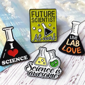Op de afbeelding: Vier emaille pins met wetenschapsthema's. Eén pin zegt "FUTURE SCIENTIST At work" op een groene achtergrond. Andere pins tonen bekers met de zinnen "I SCIENCE", "Live Lab Love" en "Science is awesome".
