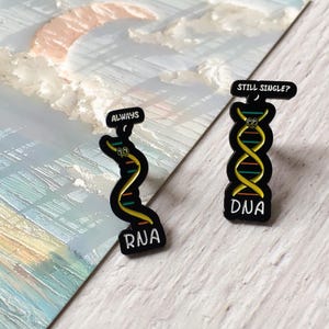 Può includere: Due spille smaltate nere a tema DNA e RNA. Una spilla recita "ALWAYS" e presenta un filamento di RNA curvo. L'altra spilla recita "STILL SINGLE?" e presenta un filamento di DNA. Entrambe le spille hanno accenti gialli, verdi e rossi.