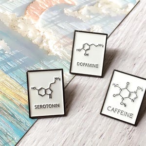 May include: Three rectangular enamel pins with black borders. Each pin features a white background with a black chemical structure and a word: Dopamine, Serotonin, and Caffeine. The pins are displayed on a surface with a watercolor design.