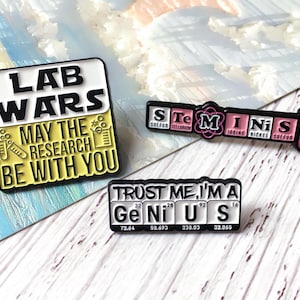 Könnte beinhalten: Emaille-Anstecknadeln mit Wissenschaftsthemen. Eine Anstecknadel sagt "LAB WARS" mit "MAY THE RESEARCH BE WITH YOU". Eine andere sagt "STEMinist". Die dritte sagt "TRUST ME, I'M A GENIUS" mit Elementsymbolen.