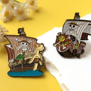 May include: Two enamel pins featuring pirate ships from One Piece. The left pin depicts the Going Merry with a sheep figurehead and a skull and crossbones sail. The right pin shows the Thousand Sunny with a lion figurehead and a sun emblem.