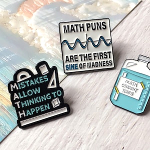 May include: Three enamel pins with math-related puns. One black pin reads "MISTAKES ALLOW THINKING TO HAPPEN." A white pin says "MATH PUNS ARE THE FIRST SINE OF MADNESS." A blue pin depicts a container labeled "MATH STUDENT TEARS."