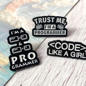 May include: Three black enamel pins with white text. One pin reads "I'M A PRO GRAMMER" with Ctrl+C and Ctrl+V symbols. Another says "TRUST ME I'M A PROGRAMMER". The third pin says "<CODE> LIKE A GIRL".