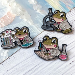 Peut inclure: Trois épingles en émail représentant des grenouilles de dessin animé en blouses de laboratoire. L'une des grenouilles utilise un microscope, une autre écrit dans un cahier et la troisième travaille avec des tubes à essai. Les épingles ont des contours noirs et sont sur un fond blanc et bleu.
