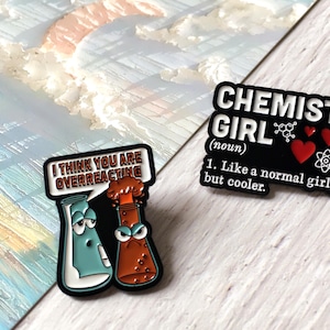 Könnte beinhalten: Zwei Emaille-Anstecknadeln mit Designs zum Thema Wissenschaft. Eine Anstecknadel zeigt zwei Cartoon-Becher mit dem Text "I THINK YOU ARE OVERREACTING". Die andere Anstecknadel sagt "CHEMIST GIRL" mit einer Definition und wissenschaftlichen Symbolen.
