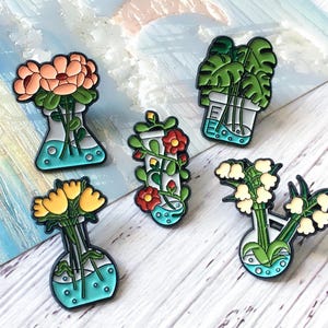May include: A collection of five enamel pins featuring flowers and plants in laboratory flasks. The pins showcase various floral arrangements with colors like peach, yellow, red, and white, set against a teal and white background.