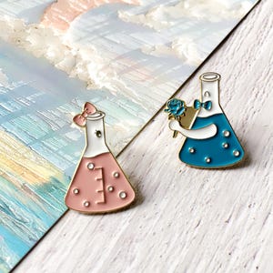 May include: Two enamel pins shaped like laboratory flasks. One is pink with a bow, the other is blue and holding a blue rose. Both have gold outlines and white accents. The pins are on a white and blue background.