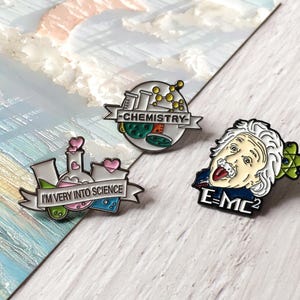 May include: Three enamel pins with science themes. One pin says "I'M VERY INTO SCIENCE" with a beaker and hearts. Another says "CHEMISTRY" with lab equipment. The third pin features Albert Einstein and the formula E=MC².