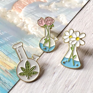 Op de afbeelding: Drie emaille pins: een bong met een cannabisblad, een fles met roze rozen en een fles met witte madeliefjes. Elke pin heeft een witte achtergrond, blauw water en gouden rand.