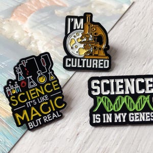 Pode incluir: Três pinos de esmalte com tema científico com bordas pretas. Um pino apresenta um microscópio e as palavras "I'M CULTURED". Outro diz "SCIENCE IS LIKE MAGIC BUT REAL". O terceiro pino diz "SCIENCE IS IN MY GENES".