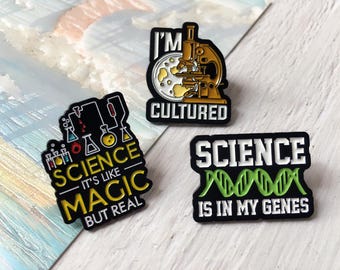 May include: Three science-themed enamel pins with black borders. One pin features a microscope and the words "I'M CULTURED." Another says "SCIENCE IS LIKE MAGIC BUT REAL." The third pin reads "SCIENCE IS IN MY GENES."