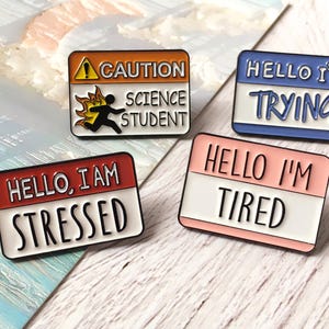 Könnte beinhalten: Vier Emaille-Anstecknadeln mit humorvollen Sprüchen. Eine Nadel lautet "CAUTION SCIENCE STUDENT" mit einer brennenden Strichmännchen-Figur. Andere Nadeln sagen "HELLO I'M TRYING", "HELLO, I AM STRESSED" und "HELLO I'M TIRED".
