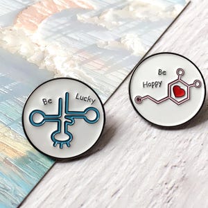 May include: Two circular enamel pins. One features a blue and white design with the words "Be Lucky". The other has a red heart and the words "Be Happy". Both pins have a black border and are on a white background.
