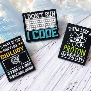 Puede incluir: Tres pines rectangulares negros con texto blanco y de colores. Uno dice "I DON'T RUN I CODE" con un gráfico de teclado. Otro dice "THINK LIKE A PROTON BE POSITIVE". El tercero dice "IT'S OKAY IF YOU DON'T LIKE BIOLOGY".