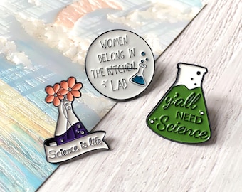May include: Three enamel pins with science-themed designs. One pin is a white circle with the text "WOMEN BELONG IN THE LAB". Another pin is a green flask with the text "y'all NEED Science". The third pin has a flask with flowers and the text "Science is life".
