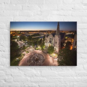 Op de afbeelding: Een luchtfoto van een grote stenen kerk met een hoge toren in de schemering. De kerk is omgeven door bomen en een geplaveide binnenplaats met een fontein in het midden. De skyline van de stad is zichtbaar op de achtergrond.