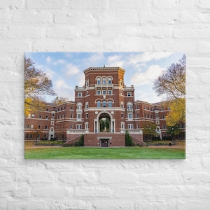 Könnte beinhalten: Leinwanddruck eines großen Backsteingebäudes mit einem zentralen Turm und einem bogenförmigen Eingang. Das Gebäude hat mehrere Fenster und ist von grünem Gras und Bäumen mit gelben Blättern umgeben. Der Himmel ist blau mit einigen Wolken.