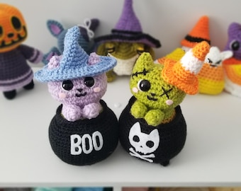 Puede incluir: Dos figuras de gato de Halloween de ganchillo en trajes de bruja y zombi, sentadas en contenedores negros en forma de caldero. El gato bruja es morado con un sombrero azul y el gato zombi es verde. Los contenedores en forma de caldero tienen texto blanco, uno dice "BOO" y el otro tiene una calavera y huesos cruzados.