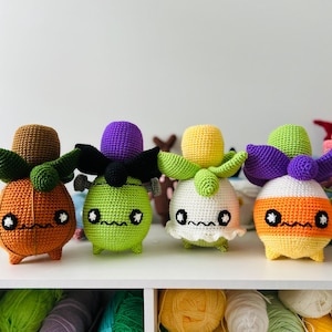 May include: Four handmade crochet dolls in Halloween colours. The dolls have round bodies, small legs, and large eyes. The dolls are orange, green, white, and orange and yellow. Each doll has a different headpiece, including a pumpkin, a witch, and candy corn.